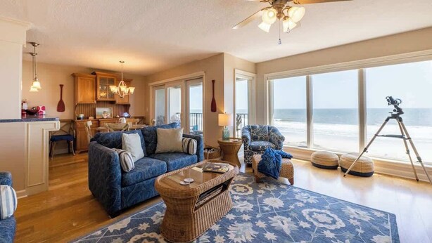 A spacious living room with a large window overlooking the beach, a ceiling fan, and a kitchen area visible through the open door. - Sur La Plage-Oceanfront & Pool (Fernandina Beach)