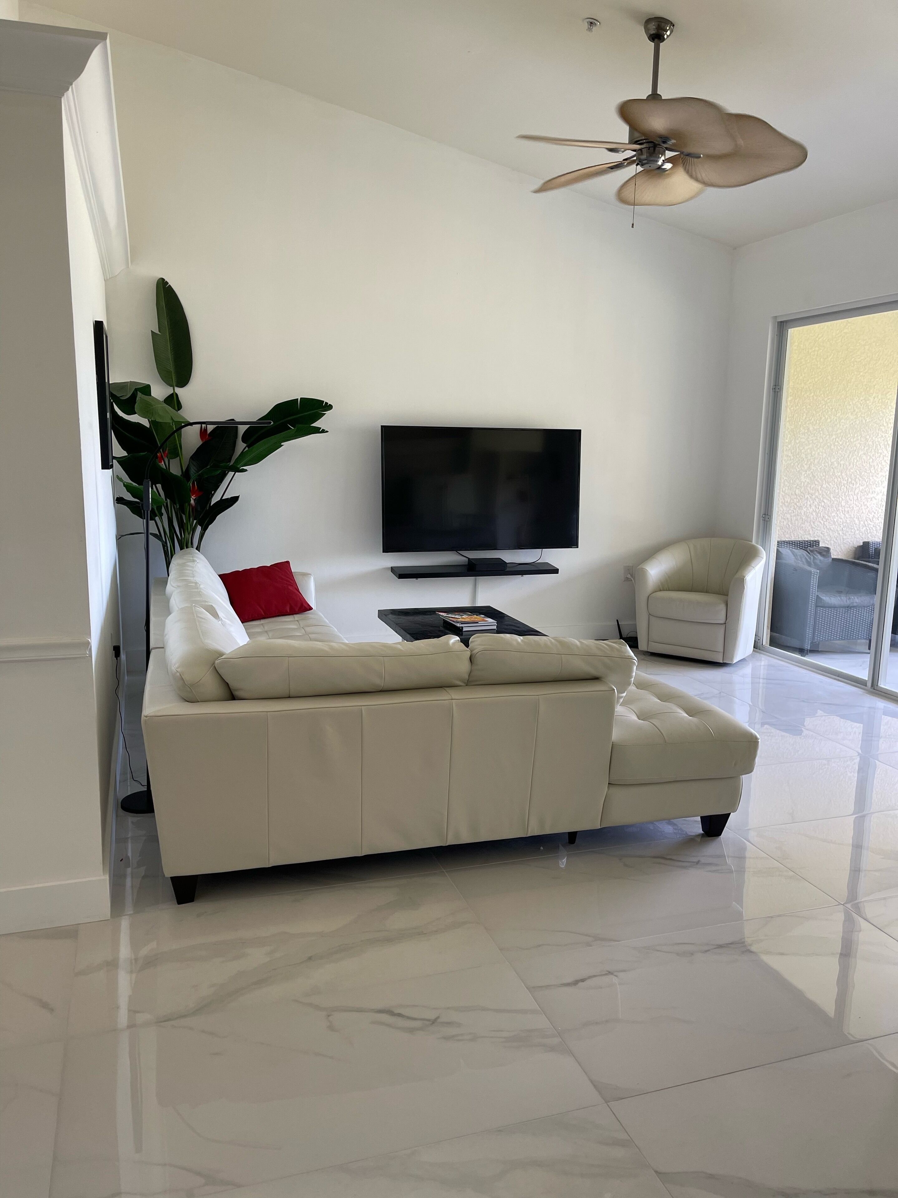 Una sala de estar moderna con un sofá de cuero blanco, un televisor de pantalla plana, un ventilador de techo y un ventanal grande.