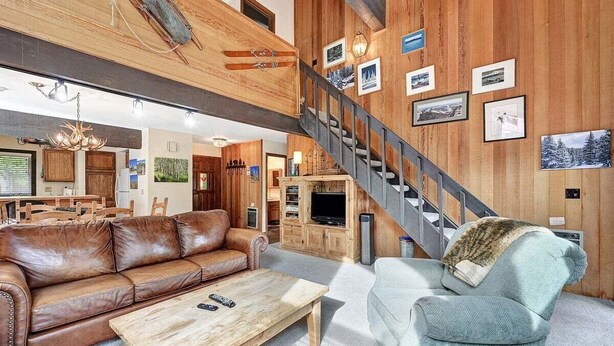 Un salón amplio con un sofá de cuero marrón, un sillón azul claro y una mesa de centro de madera. Una escalera con barandilla de metal conduce a otro nivel.