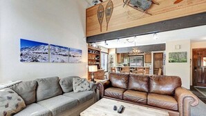 Una sala de estar con un sofá, una mesa de centro y una chimenea. Se observan muebles de madera y una cocina al fondo.