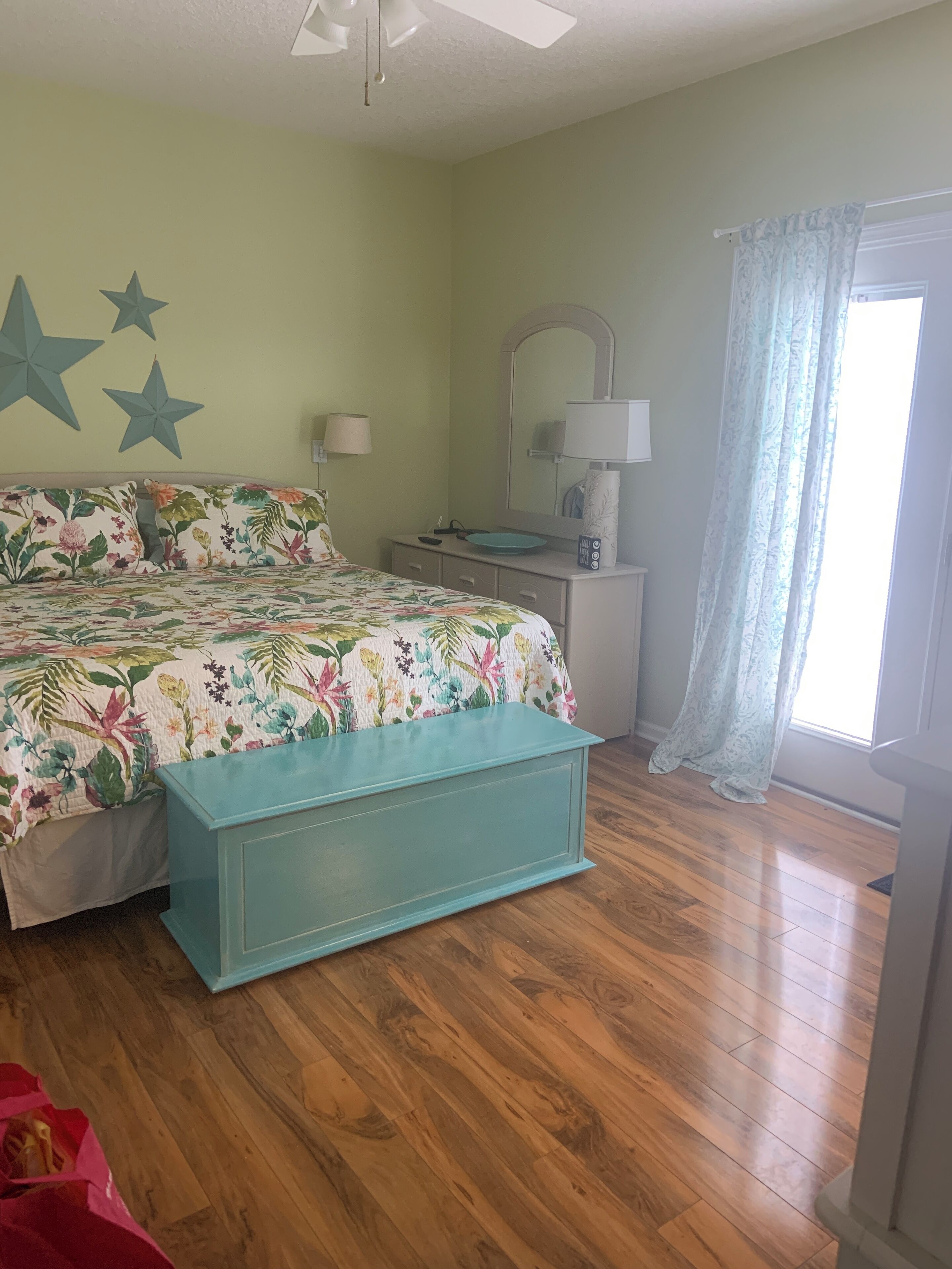 A bedroom with a floral bedspread, a teal bench, a nightstand with a lamp, and a window with a sheer curtain.