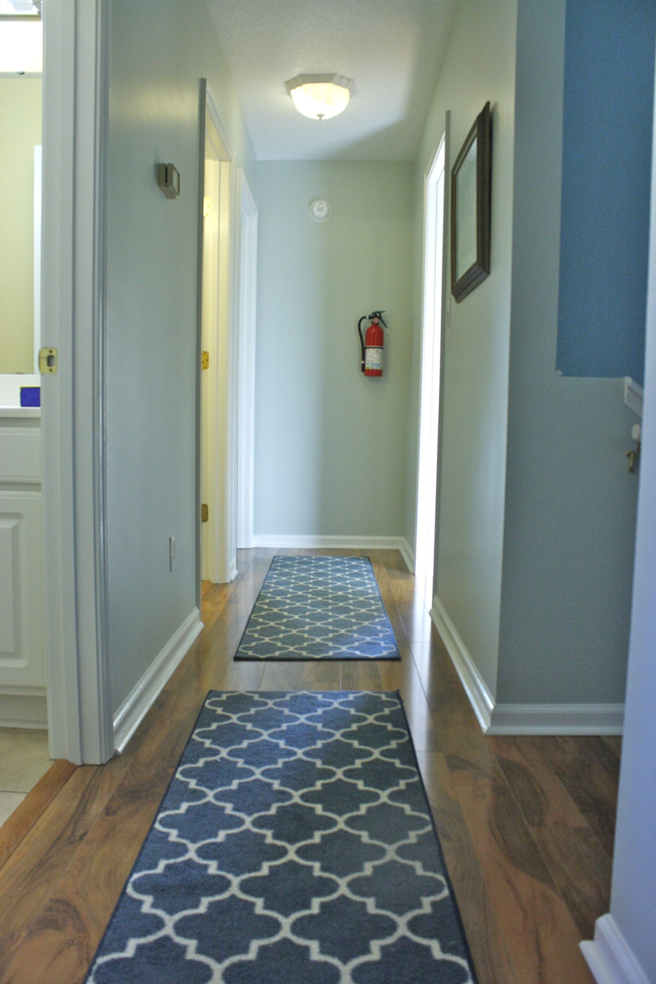 A hallway with wooden floors, a patterned rug, a fire extinguisher, and a door leading to a room.