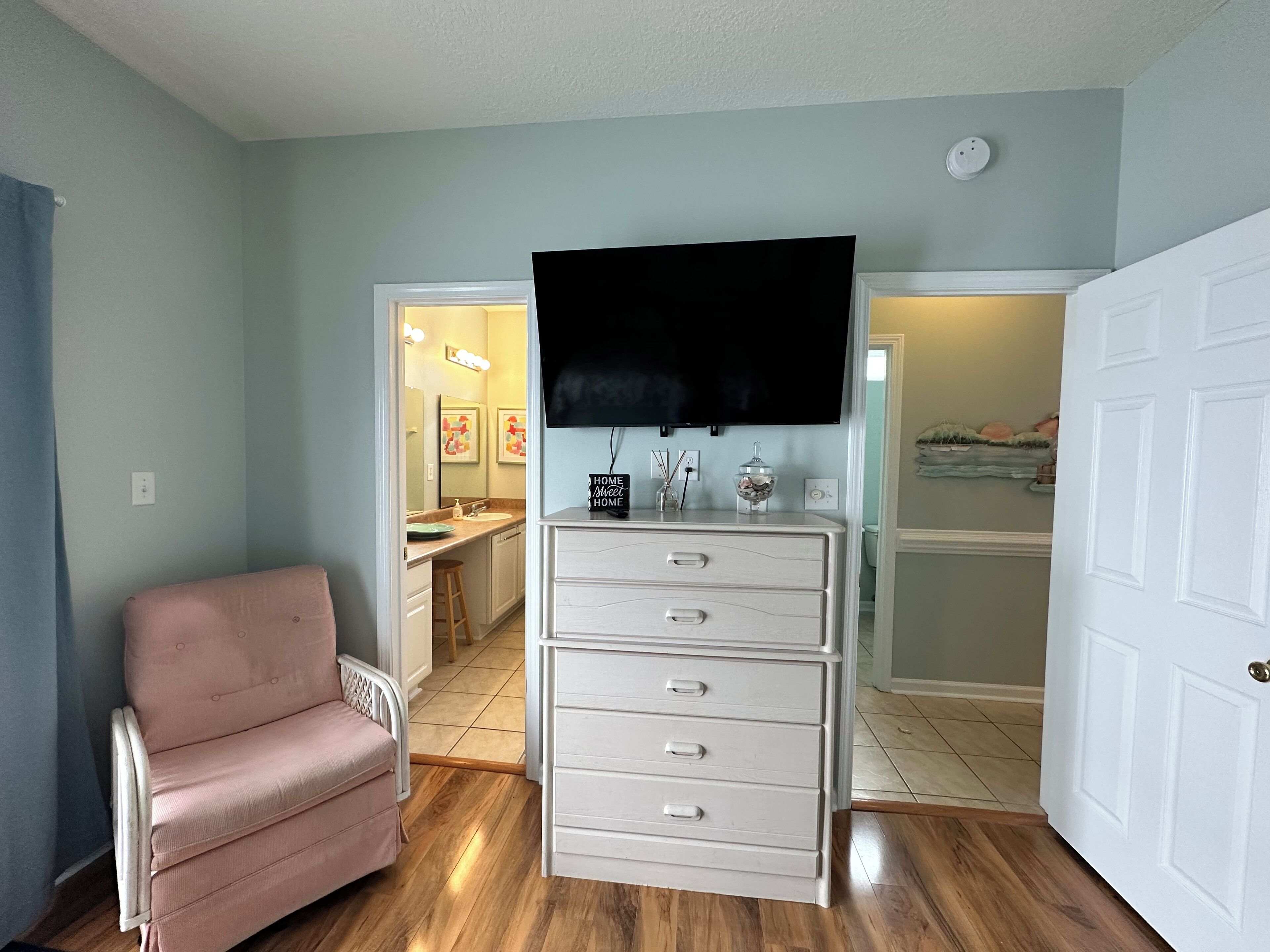 A living room with a pink armchair, a white dresser with drawers, a flat-screen TV mounted above it, and a door leading to another room.