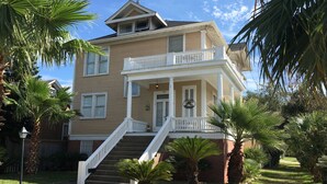 A two-story house with a front porch, a staircase, and a small front yard with palm trees.