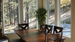 Un coin repas avec une table et des chaises en bois, une plante en pot et de grandes fenêtres donnant sur une zone boisée.