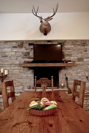 A dining area with a stone fireplace, a mounted deer head, and a wooden table set with fruits and vegetables.