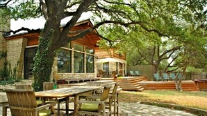 A patio area with a stone table and chairs, a wooden pergola, and a stone building with large windows. - Austin Lake Travis Pool Waterfront -  Hill Country Retreat 2 Acres  Sleeps 20 (Leander)