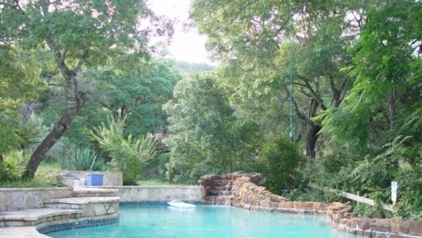 A swimming pool with a stone edge, surrounded by lush greenery and trees. - Austin Lake Travis Pool Waterfront -  Hill Country Retreat 2 Acres  Sleeps 20 (Leander)