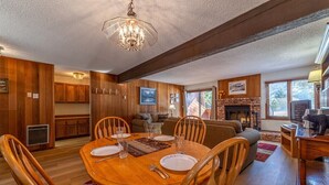Uma sala de jantar com uma mesa de madeira posta para quatro pessoas, rodeada por cadeiras de madeira. Há uma lareira com revestimento de pedra, um sofá e uma televisão ao fundo.