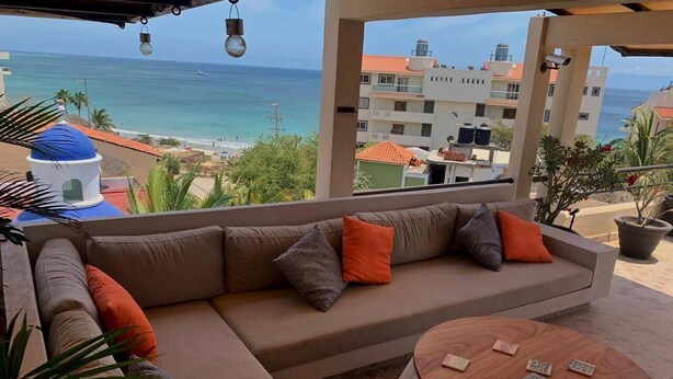 Un espace extérieur aménagé, abrité et doté d’un canapé et d’une table ronde, offrant une vue sur une plage et des bâtiments.
