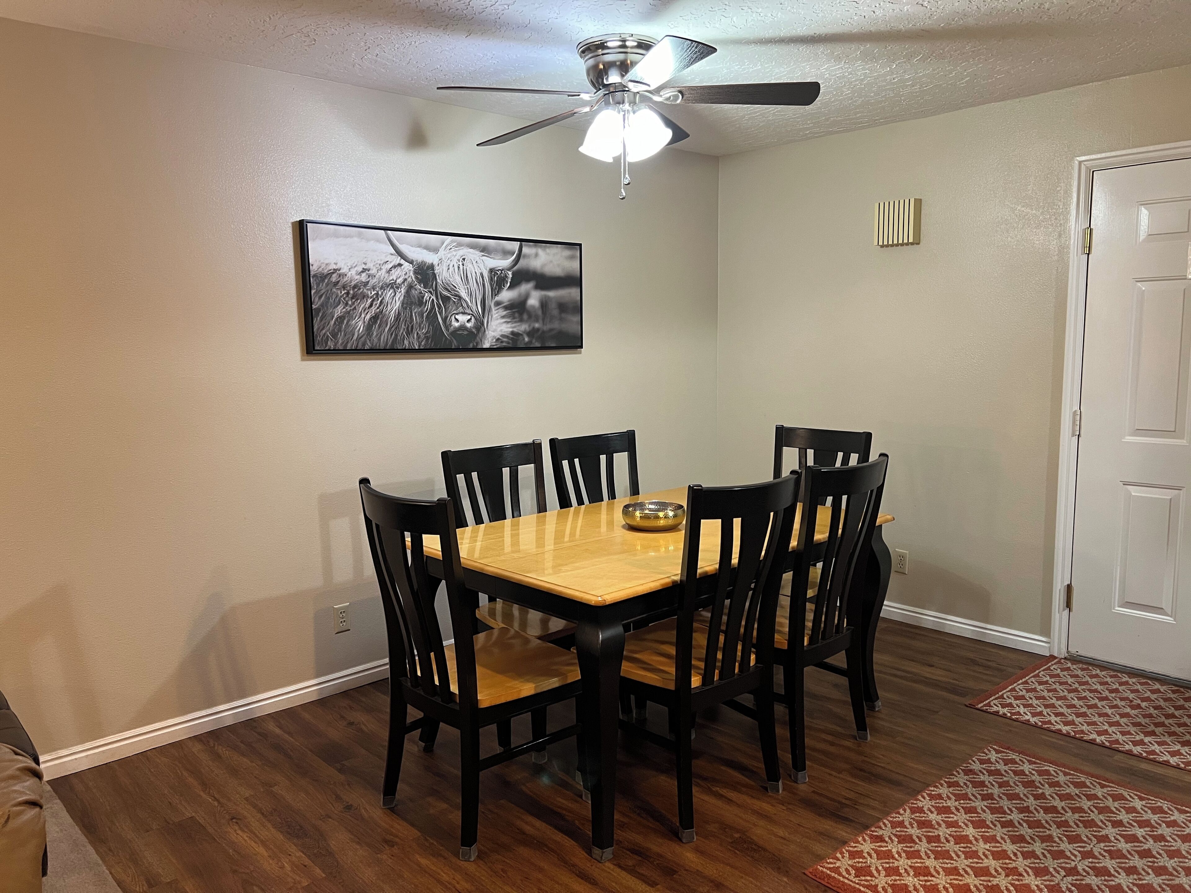 A dining room with a wooden table and black chairs, a ceiling fan, a framed picture on the wall, and a door with a doormat.