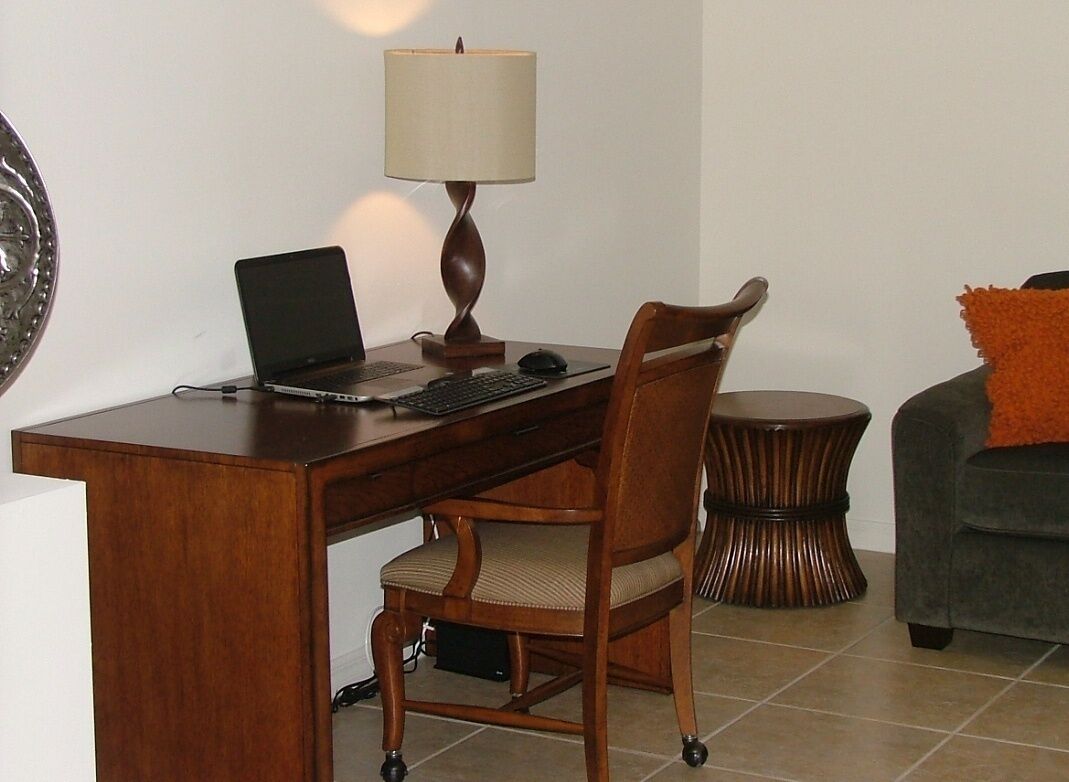 A wooden desk with a laptop, a lamp, and a chair in a room with a tiled floor and a round side table.