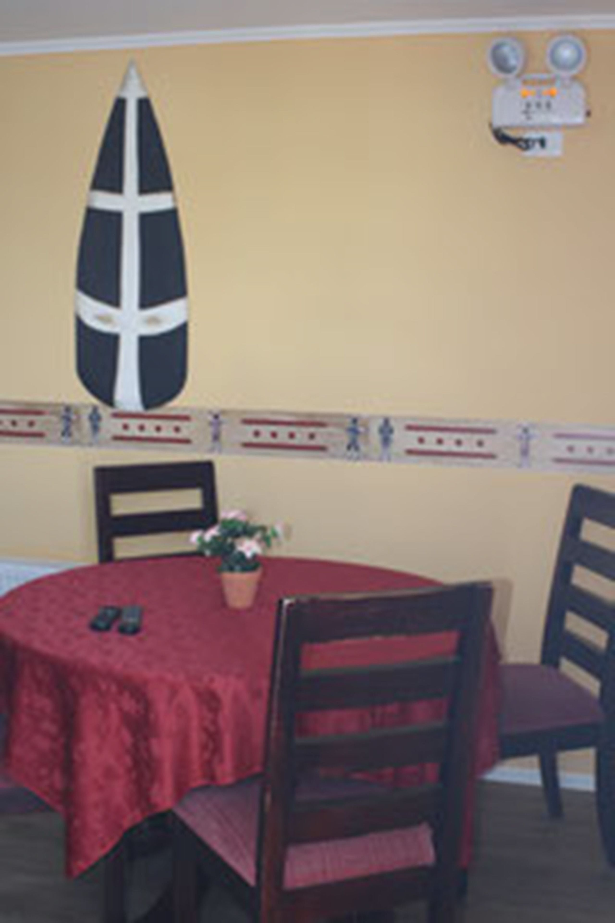 A dining area with a round table covered in a red tablecloth, two chairs, a potted plant, and a wall-mounted light fixture.