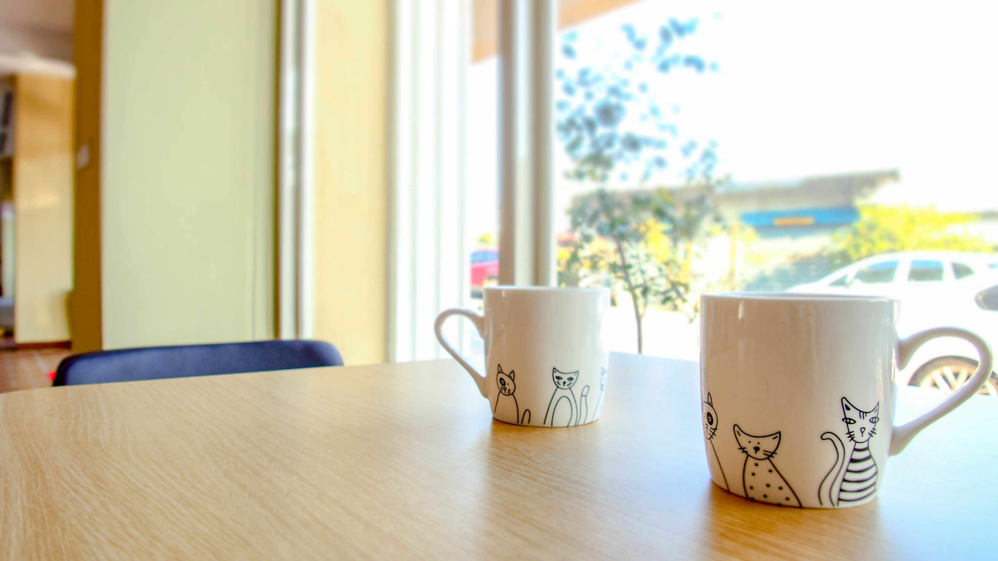 Two white mugs with cat illustrations on a wooden table near a window.