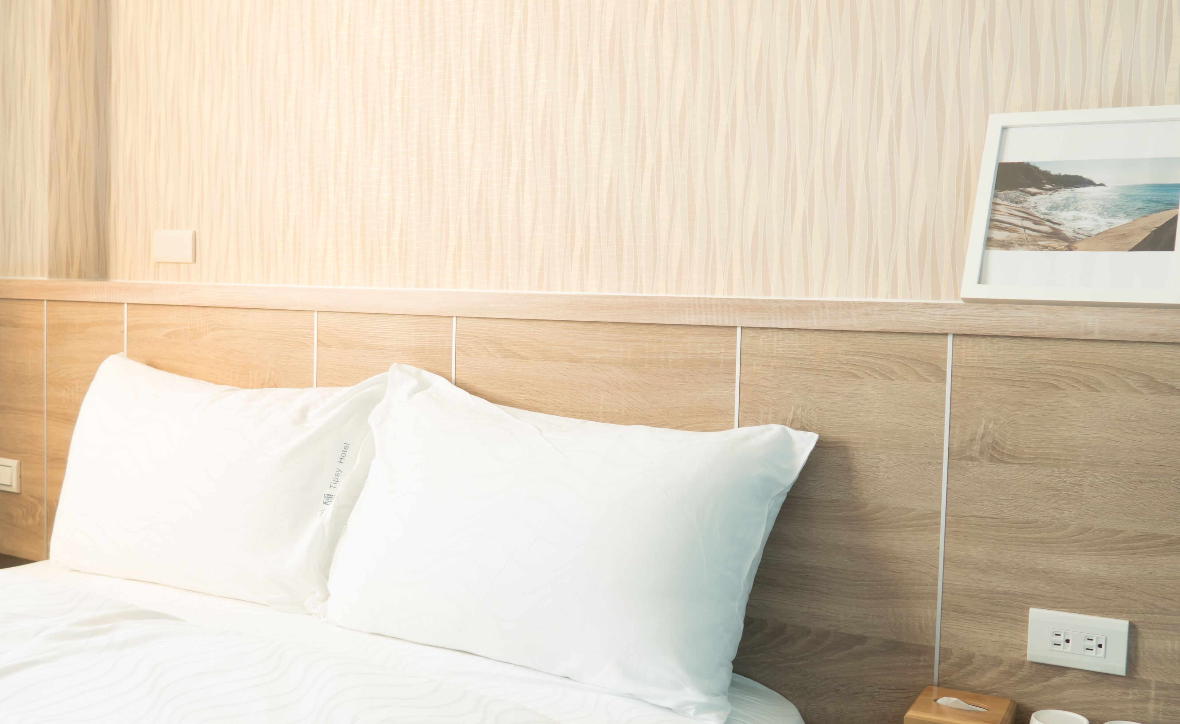 A hotel room with a wooden headboard, white bedding, and a framed picture of a beach on the wall.