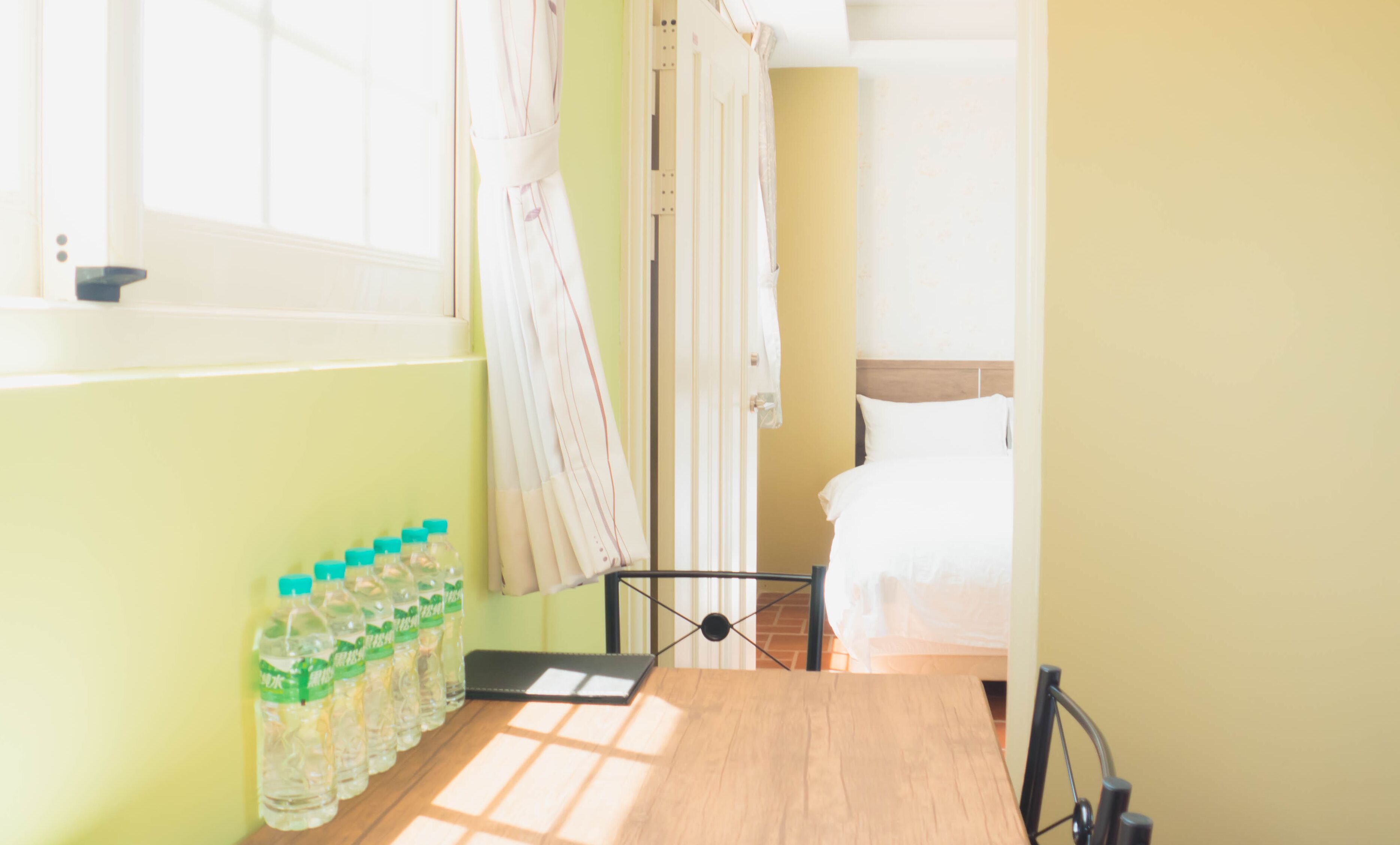 Une pièce avec un lit, une fenêtre avec des rideaux et une table sur laquelle se trouvent des bouteilles d’eau.