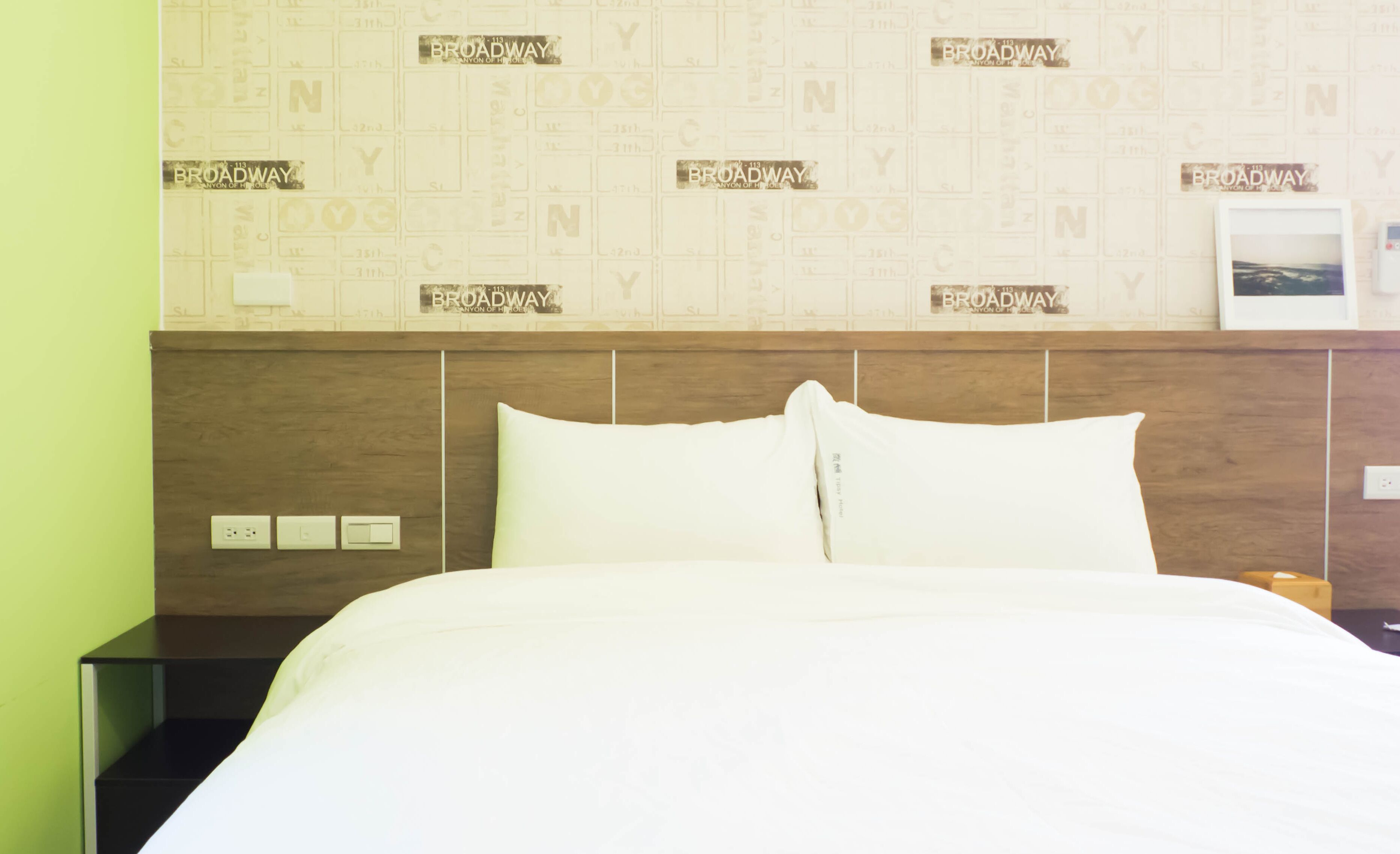 Una habitación de hotel con una cama, cabecera de madera y una pared que repite la palabra 'Broadway'.