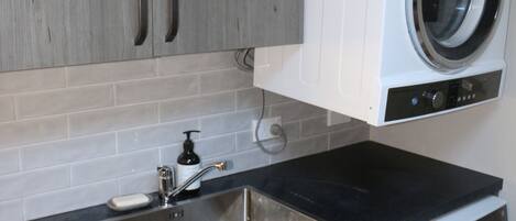 A compact kitchen with grey cabinets, a black countertop, a stainless steel sink, and a built-in washing machine.