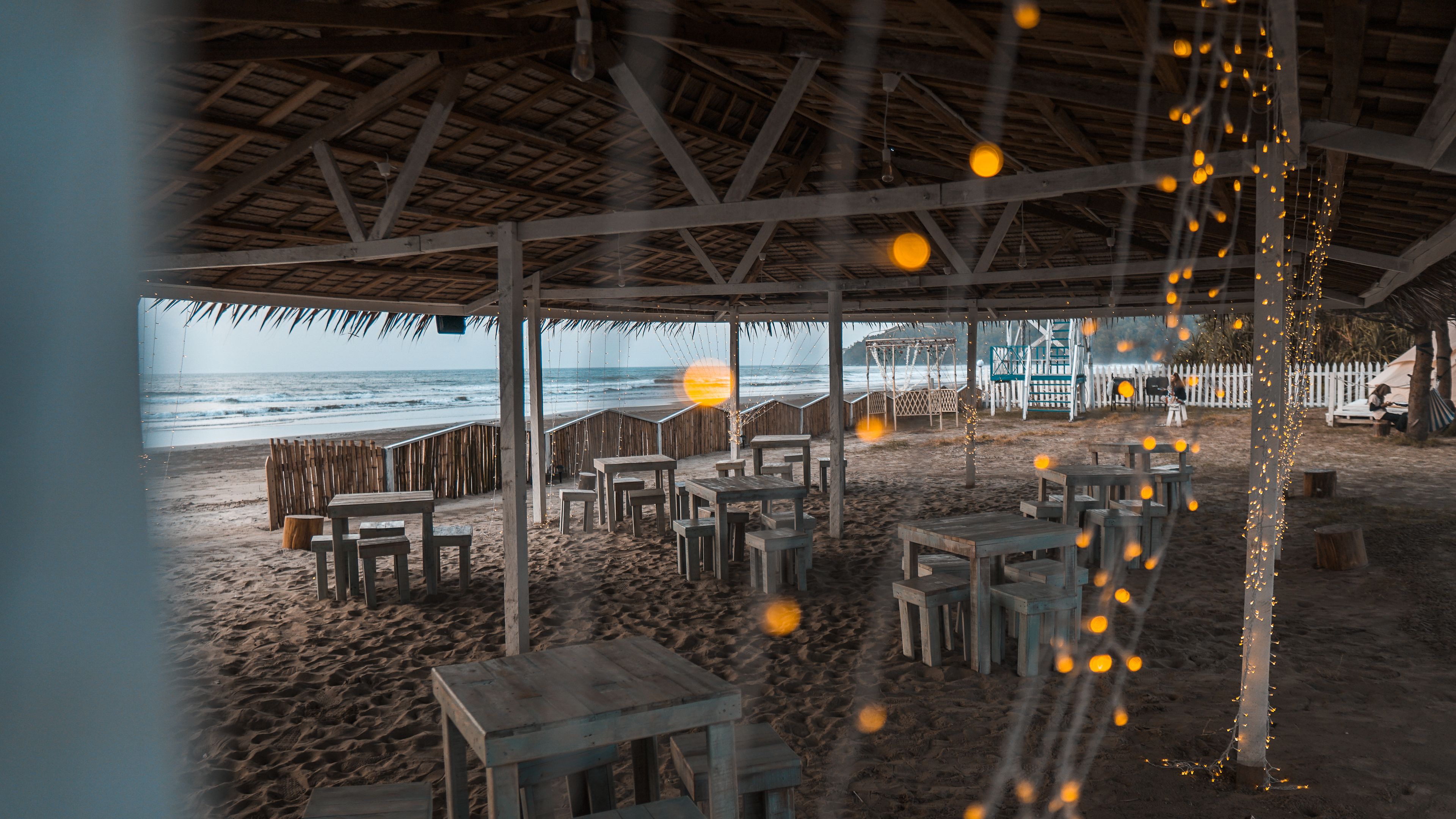 An outdoor seating area with wooden tables and chairs, a thatched roof, and a view of the beach and ocean.