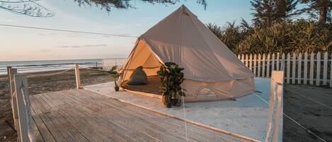 A beach tent on a wooden deck with a white picket fence, surrounded by sand and vegetation.