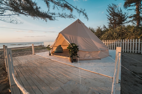 Una carpa de playa en una terraza de madera con cerca blanca, rodeada de arena y vegetación.