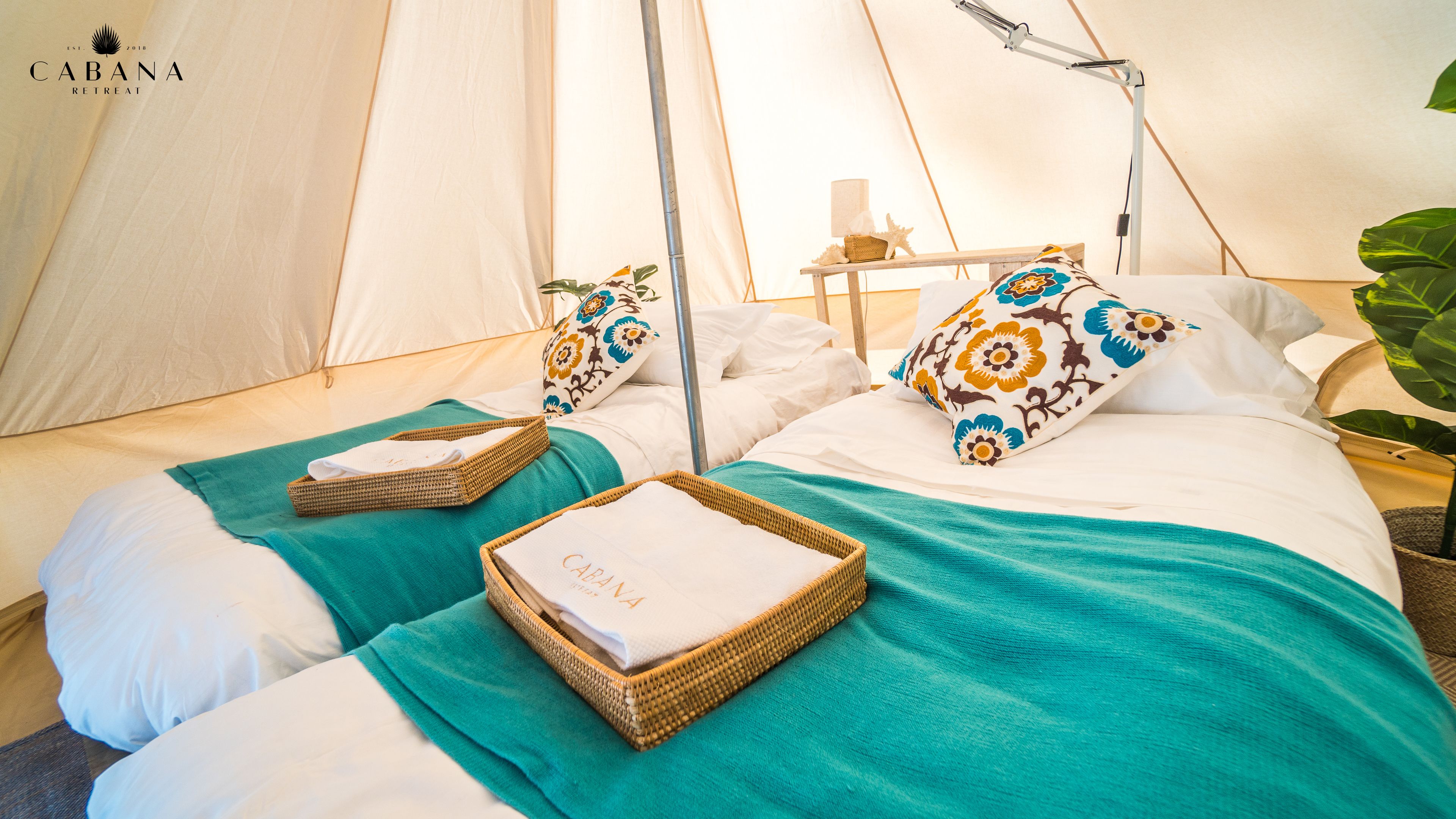 Una tienda acogedora y bien equipada con dos camas, cada una con una manta color turquesa, ropa de cama blanca y almohadas decorativas. Hay cestas de mimbre sobre las camas y una mesita con una lámpara y objetos decorativos.