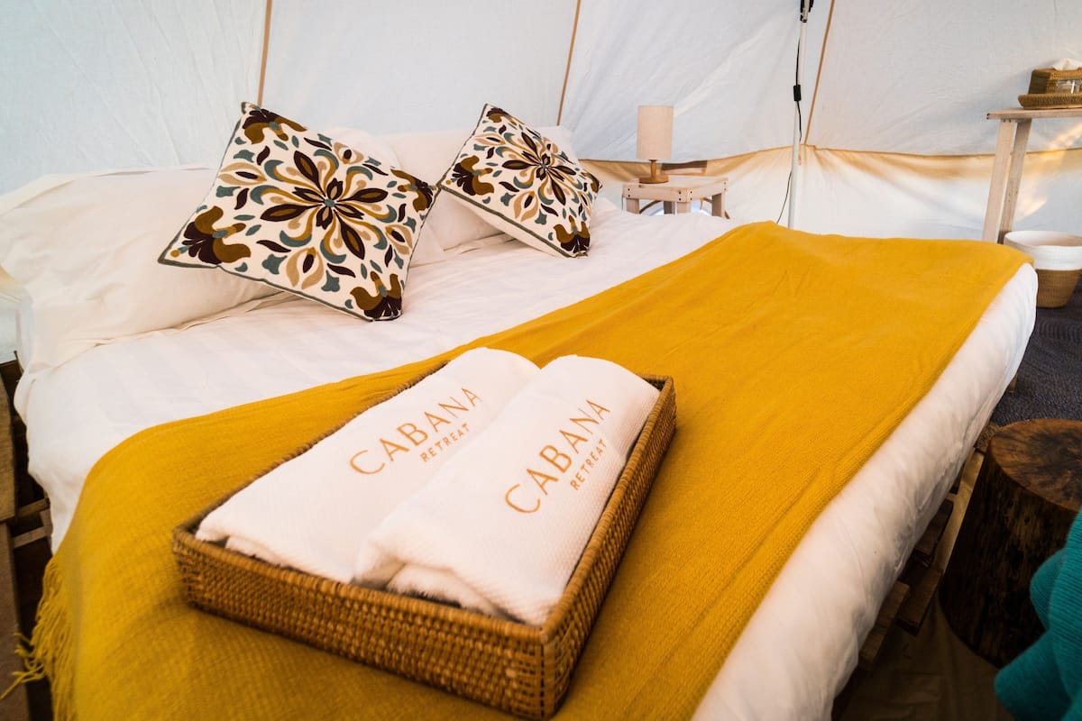 Sängyssä on keltainen peitto, kaksi kuviollista tyynyä ja kori, jossa on pyyhe, ja korissa on kyltti, jossa lukee: ”CABANA RETREAT”.