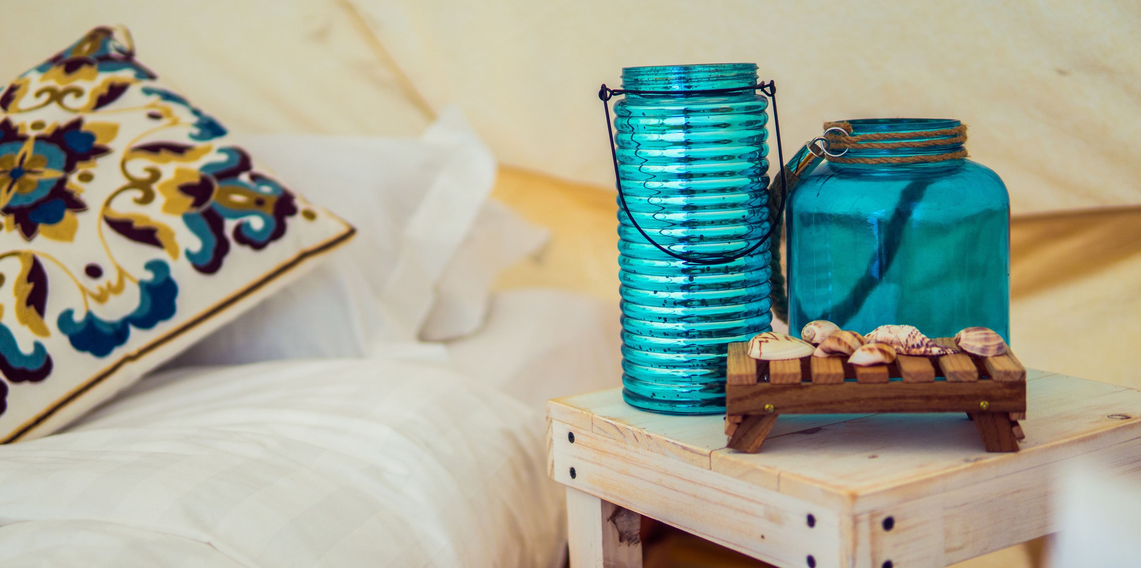 Une table de chevet en bois, un bocal en verre turquoise et un plus petit bocal, une caisse en bois remplie de coquillages et un coussin décoratif à motifs floraux.