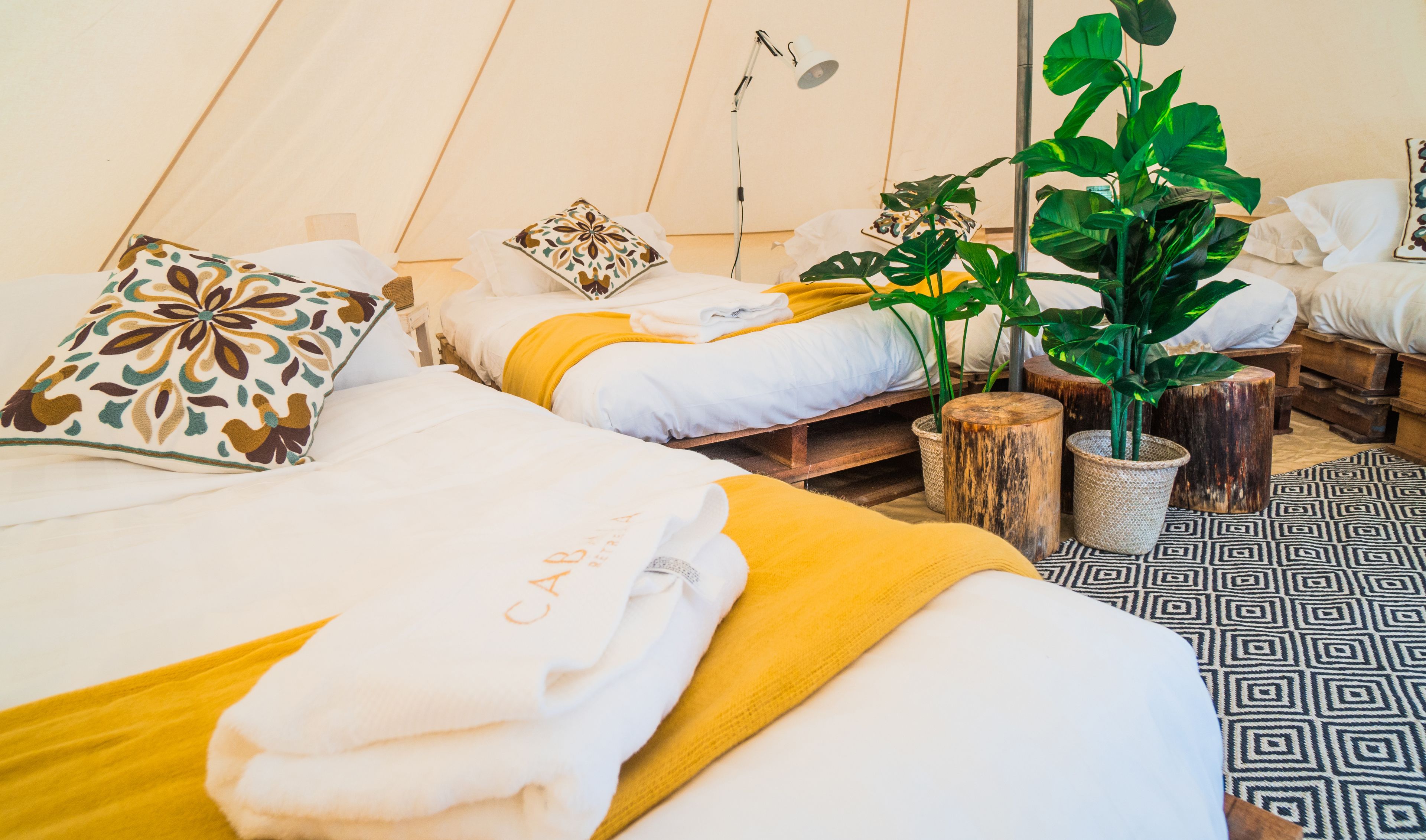A cozy, well-lit tent-style room with multiple beds, each with white bedding and decorative pillows. There are potted plants and wooden stools, creating a warm and inviting atmosphere.