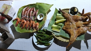 A table with a variety of grilled meats and vegetables, including sausages, shrimp, and a whole roasted chicken, served on banana leaves.