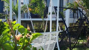A white metal swing chair with a blue roof, surrounded by greenery and a building with a blue roof and white walls in the background.