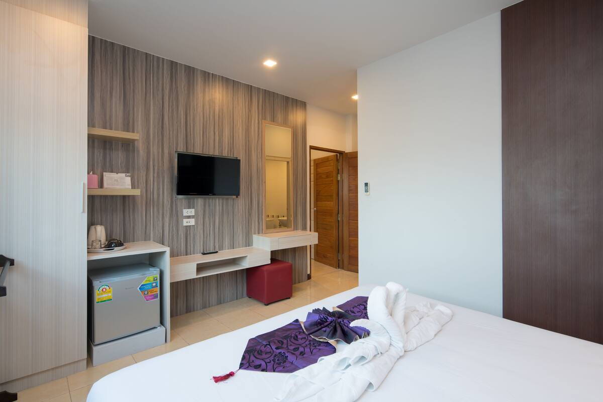 Habitación de hotel con cama, televisión, refrigerador pequeño, taburete rojo y armario de madera.