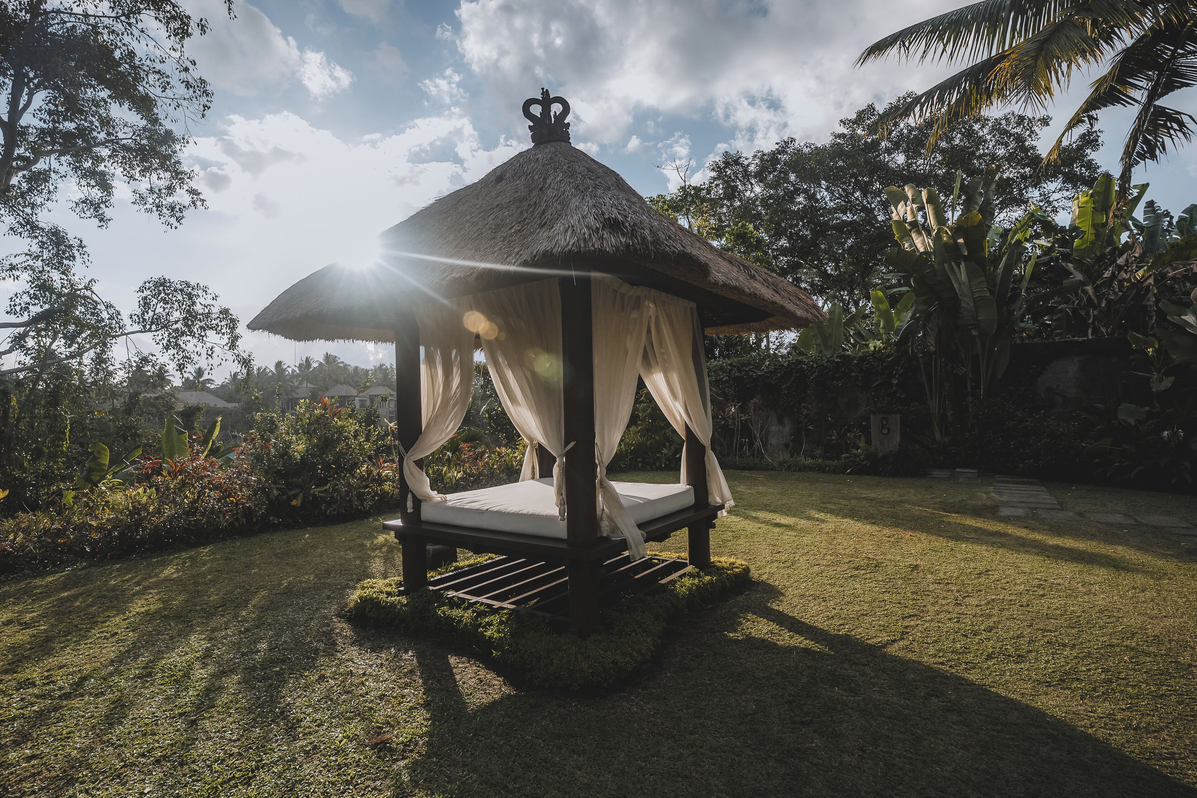 A thatched-roof gazebo with a bed inside, surrounded by lush greenery and a clear sky.
