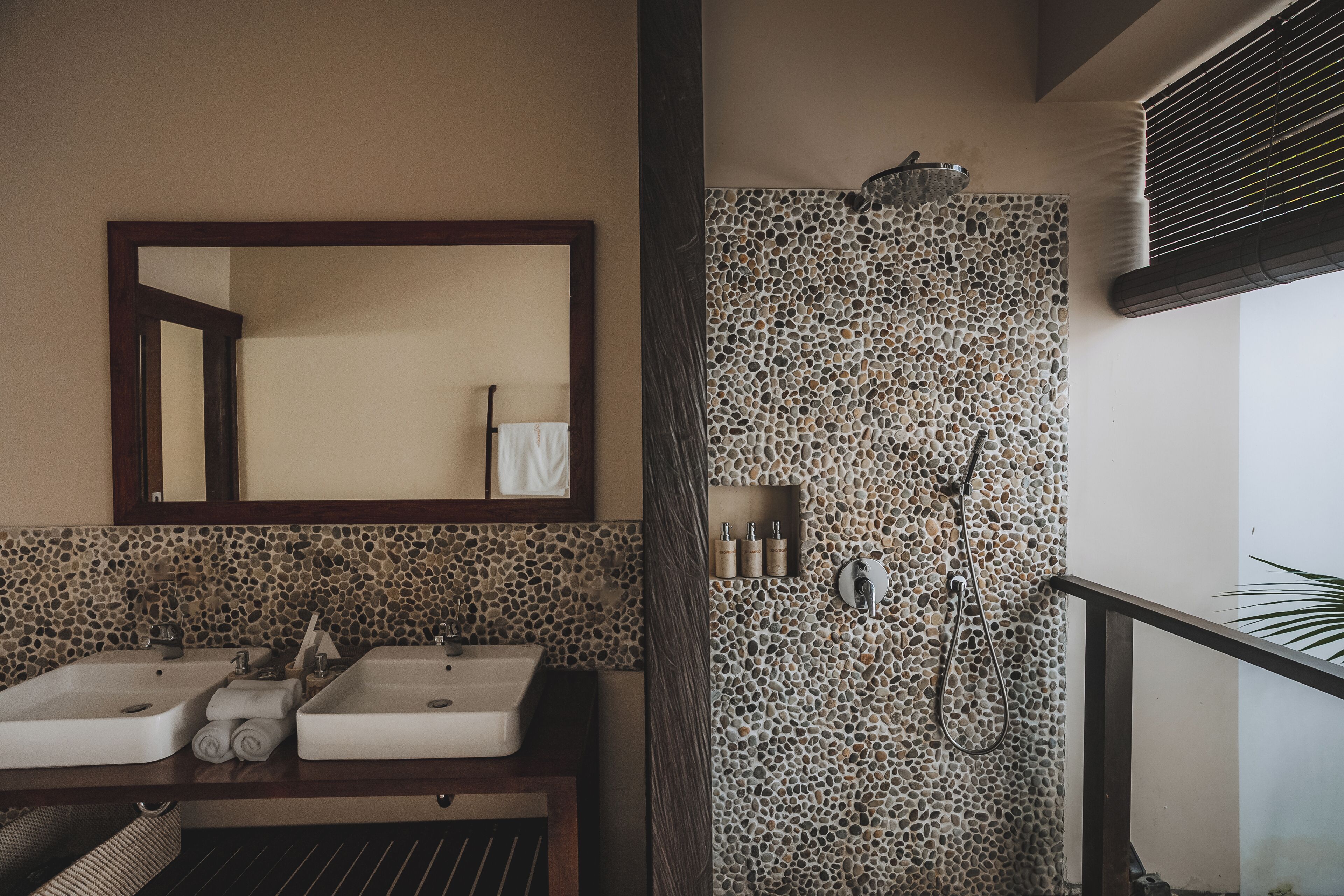 Une salle de bain avec deux lavabos, un miroir, une douche avec un mur en pierre et un porte-serviettes.