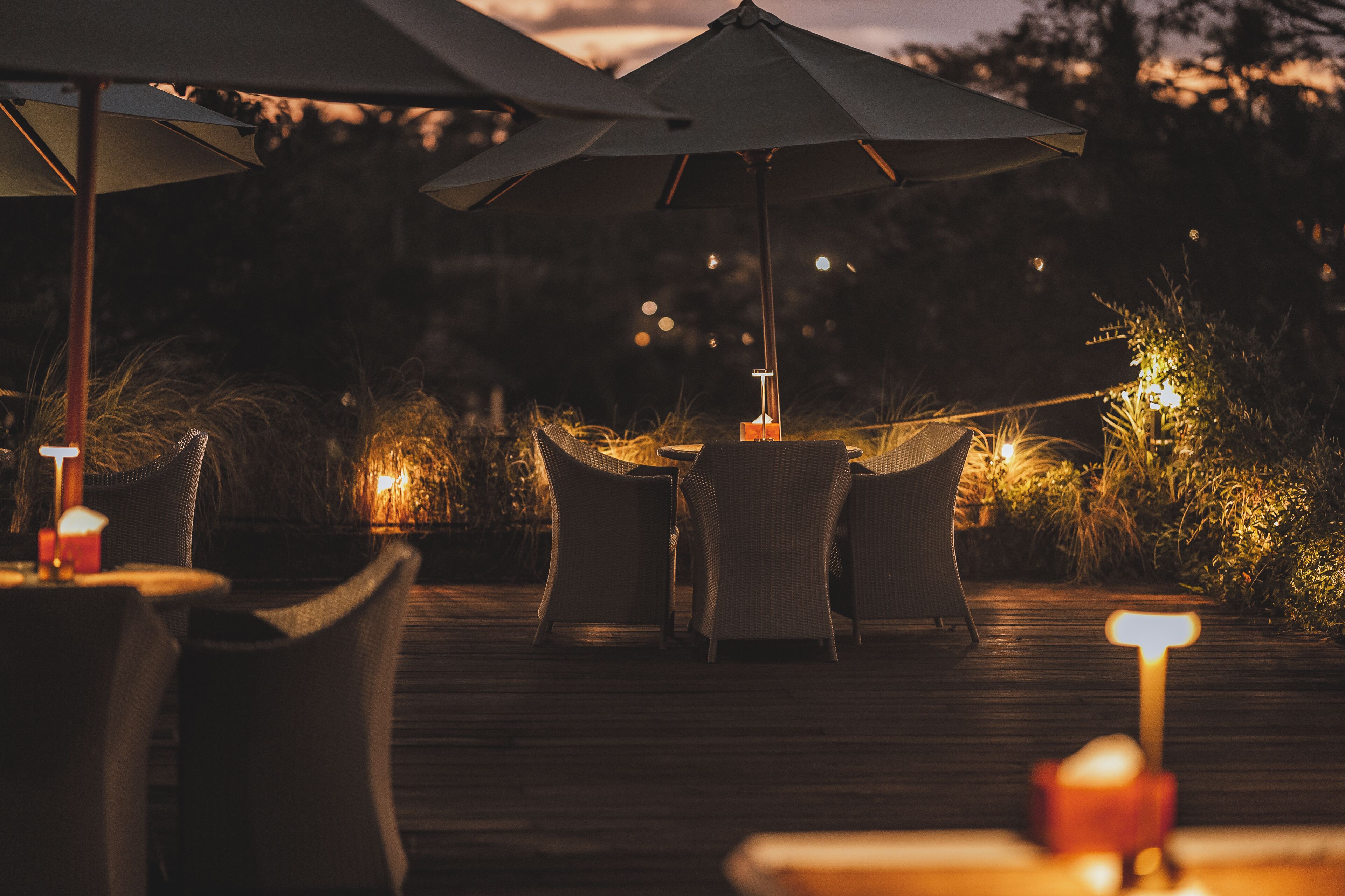 Un área de comedor al aire libre con mobiliario de mimbre y sombrillas, que cuenta con una terraza de madera e iluminación ambiental.