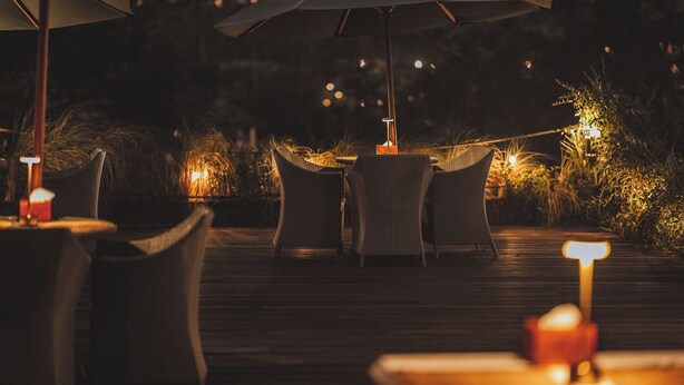 Un espace de restauration en plein air doté de mobilier en osier et de parasols, avec une terrasse en bois et un éclairage d’ambiance.