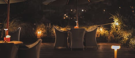 An outdoor dining area with wicker furniture and umbrellas, featuring a wooden deck and ambient lighting.