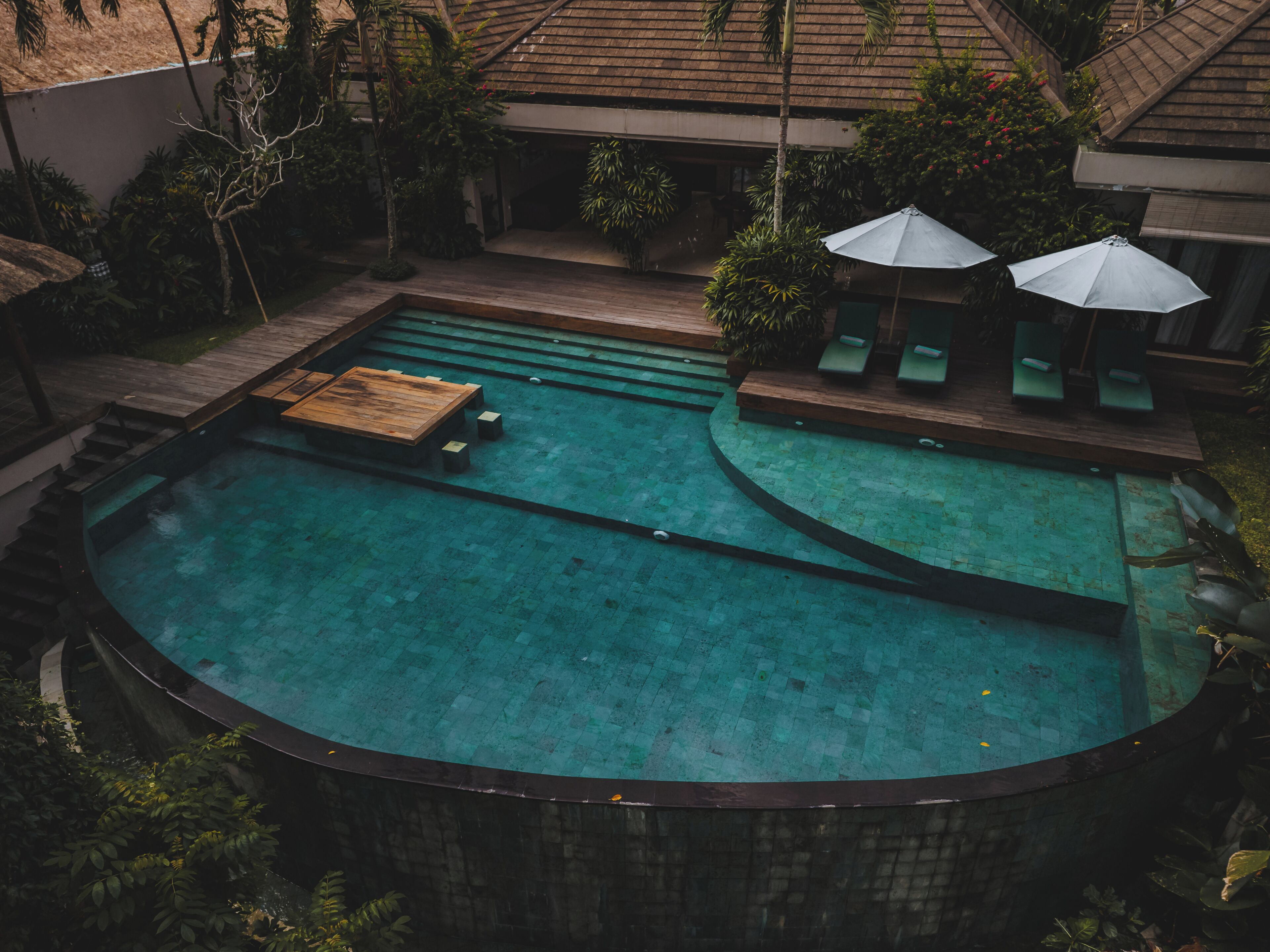 A circular swimming pool with a wooden deck and lounge chairs, surrounded by tropical plants and a covered seating area.