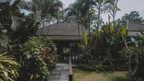 Un sentier en bois mène à un bâtiment doté d’un toit en pente, qui est entouré d’une végétation tropicale luxuriante.