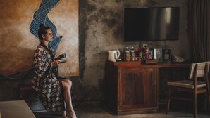 Une personne est assise sur un canapé dans une pièce où l’on aperçoit un grand téléviseur à écran plat fixé au mur, un meuble en bois contenant divers objets et un motif décoratif sur le mur.