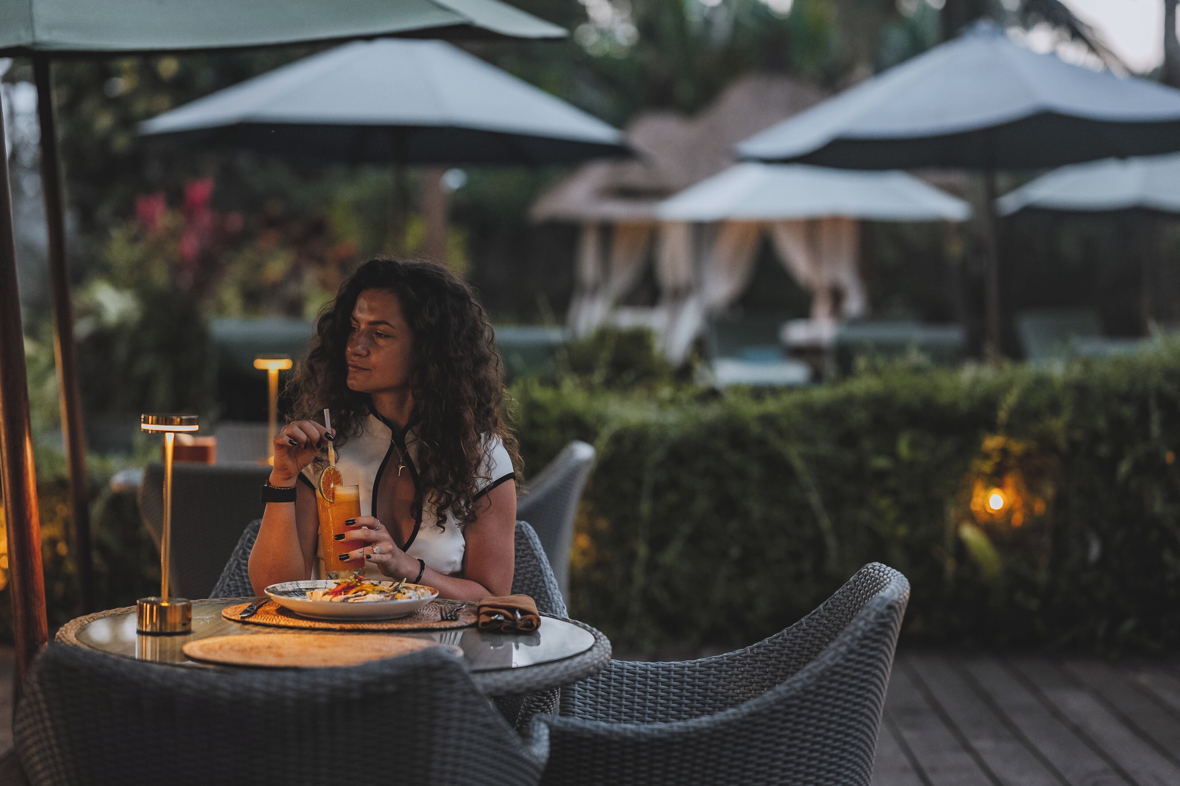 A person seated at an outdoor table with a drink, surrounded by umbrellas and greenery.