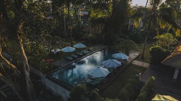 Un área de piscina con sillas de descanso y sombrillas, rodeada de árboles y un edificio con techo de paja.