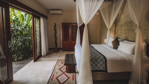A bedroom with a bed, a bench, a wooden wardrobe, and a view of greenery through a glass door.