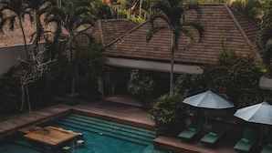 A swimming pool with a wooden deck, surrounded by lounge chairs and umbrellas, with a building and tropical vegetation in the background.