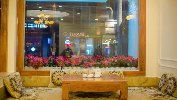 A seating area with a wooden table and chairs, a window with a view of a shopping center, and a decorative plant. - Hong Tung Hotel (Xuan Huong - Da Lat)