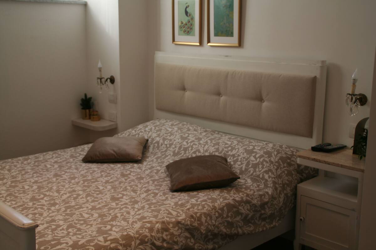 Um quarto com uma cama, duas almofadas, uma mesa de cabeceira, uma luminária e duas molduras na parede.