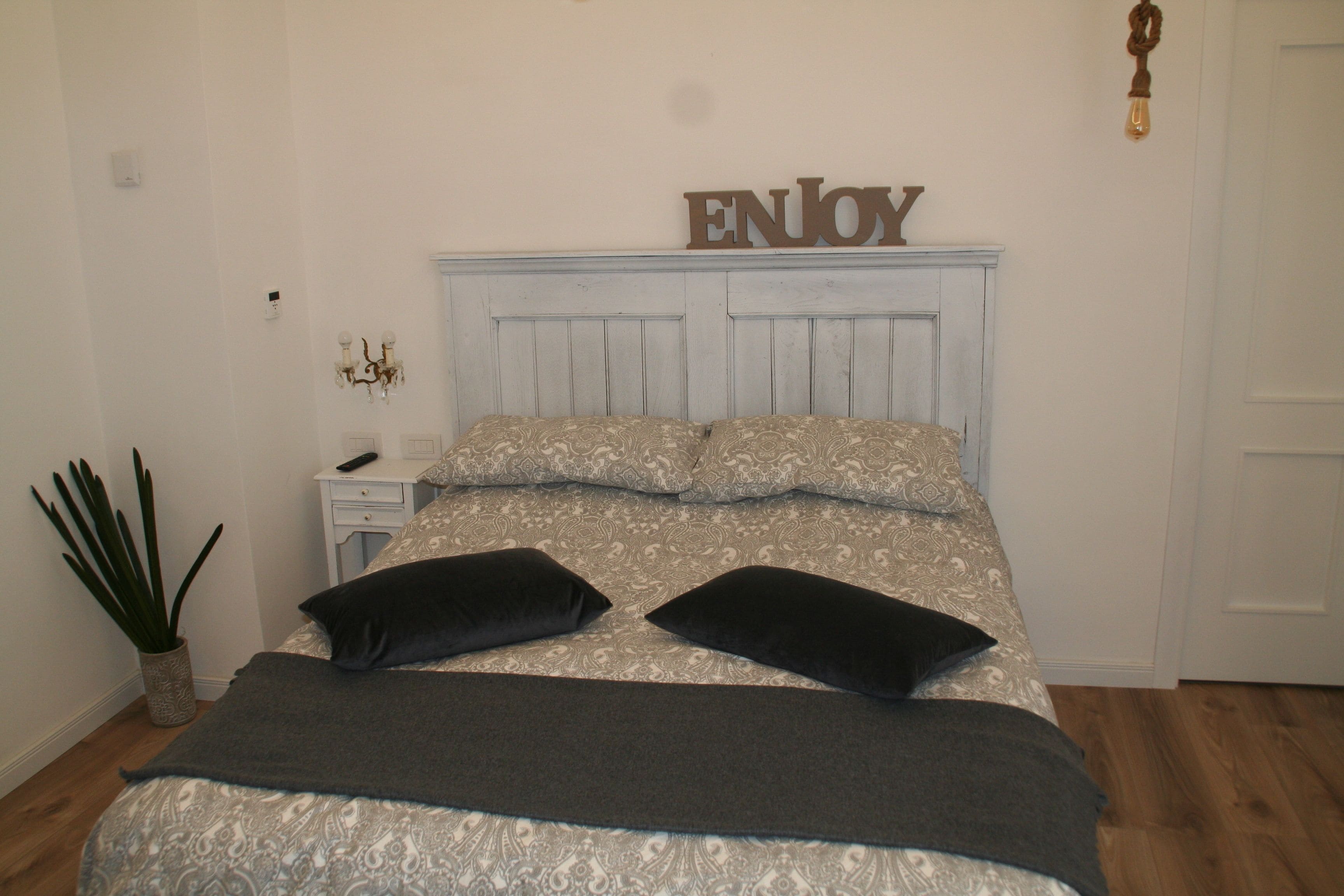 A bedroom with a bed, a nightstand, a potted plant, and a wall sign that says "ENJOY".