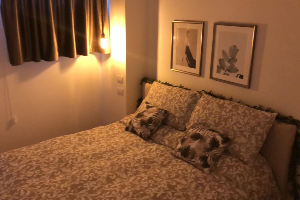 Quarto com colcha estampada, duas almofadas, cabeceira com folhagem e duas imagens emolduradas na parede.