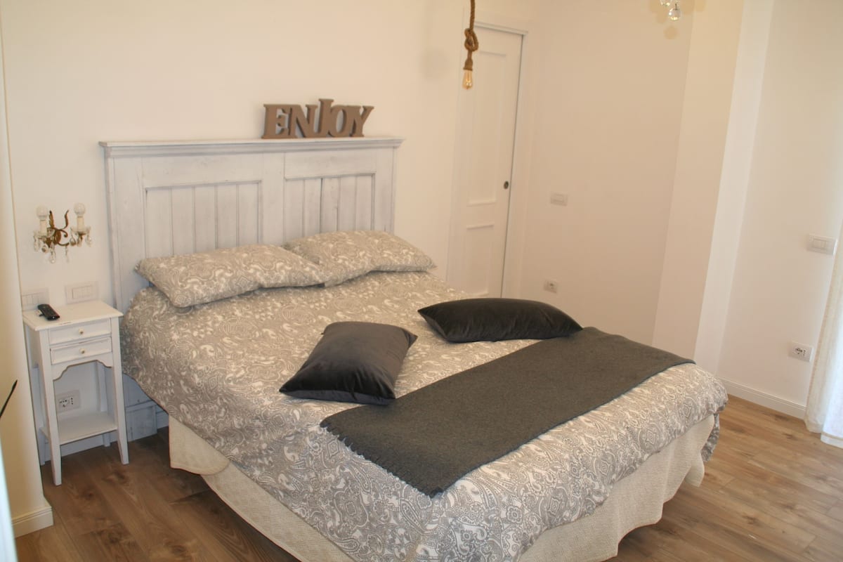 Quarto com cama, mesa de cabeceira, luminária na parede e uma porta.