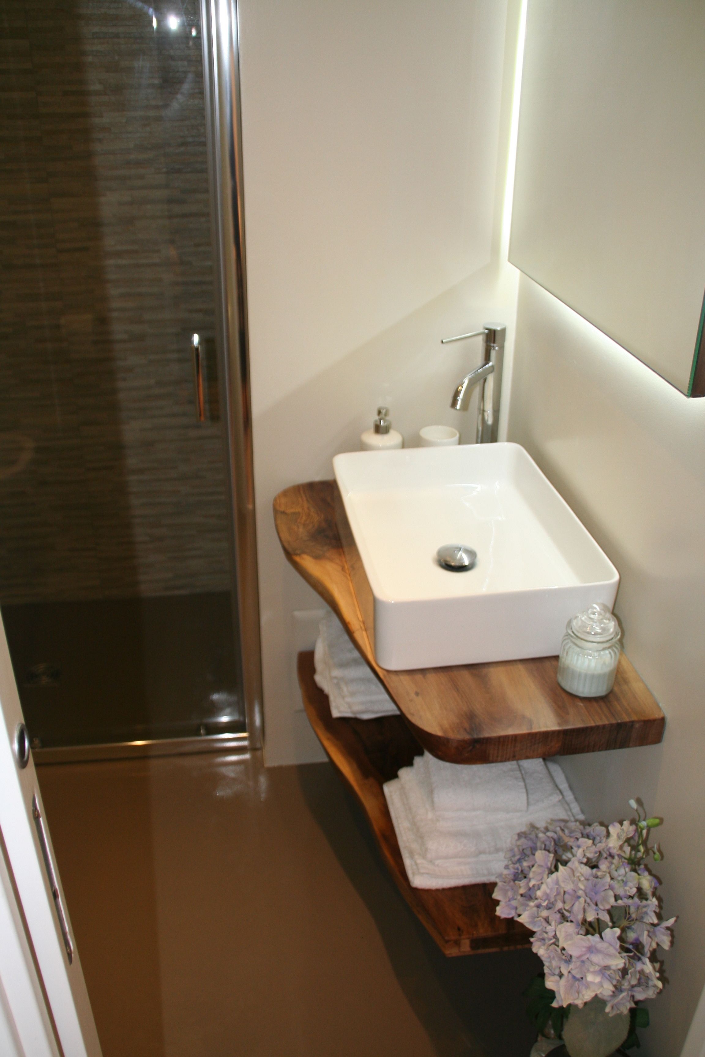 Un baño con mampara de vidrio, un lavamanos blanco con estante de madera y un jarrón con flores.